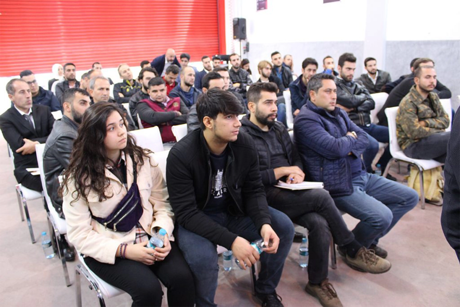 A group of people sitting attentively on chairs in a room with red shutters. Some are taking notes while others are listening. The atmosphere suggests a seminar or workshop setting.