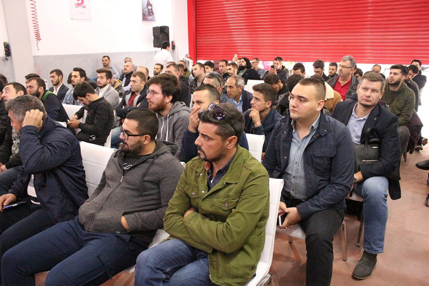A diverse group of people, mostly men, are seated in rows, attentively facing the front in a seminar or meeting room. The background features a red shutter and some posters on the wall, adding color to the scene.