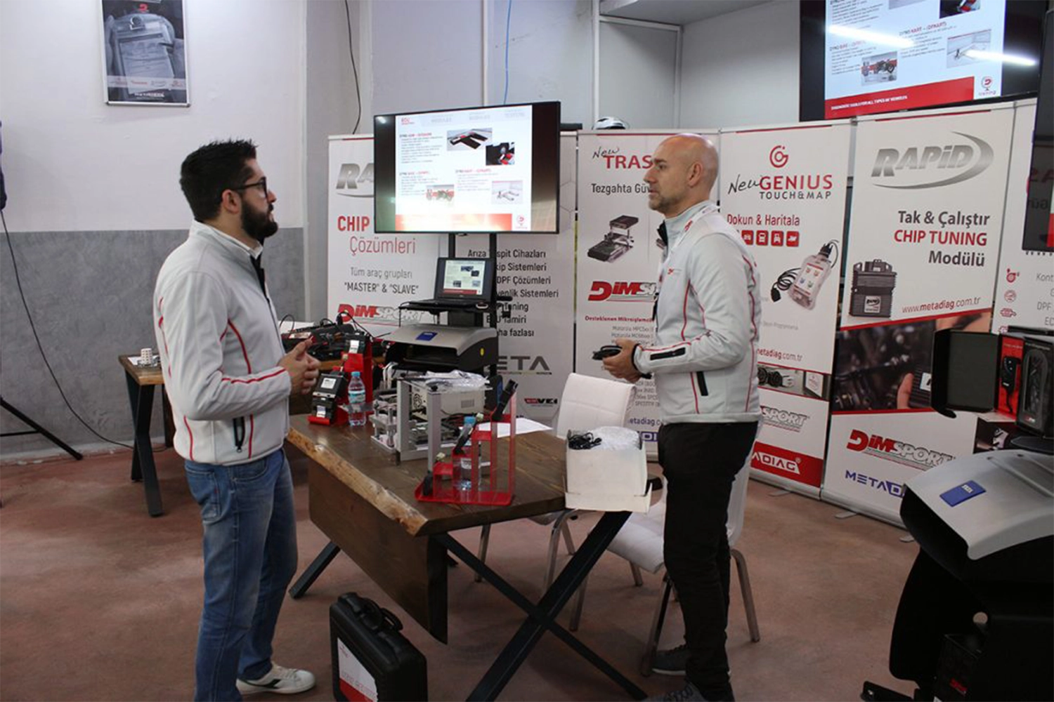 Two men are standing at a trade show booth, discussing chip tuning technology. The booth displays equipment, signage, and informational screens. The men are wearing white jackets with red accents.