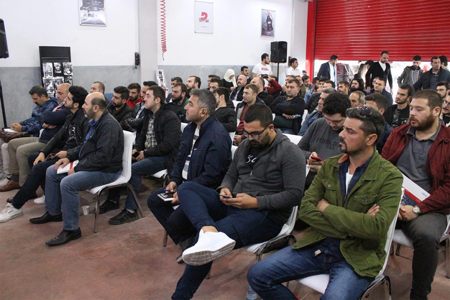 A group of people seated in rows facing forward in a large room, attentively listening to a presentation. Several individuals are taking notes or using electronic devices. The room has posters, red walls, and a raised garage door.